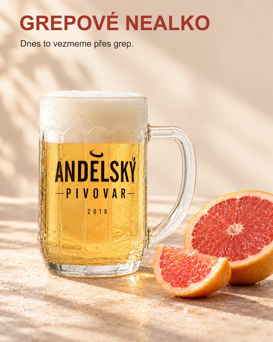 Grepové nealko ve fazetovaném půllitru Andělského pivovaru vedle rozkrojeného růžového grepu na světlém podkladu.