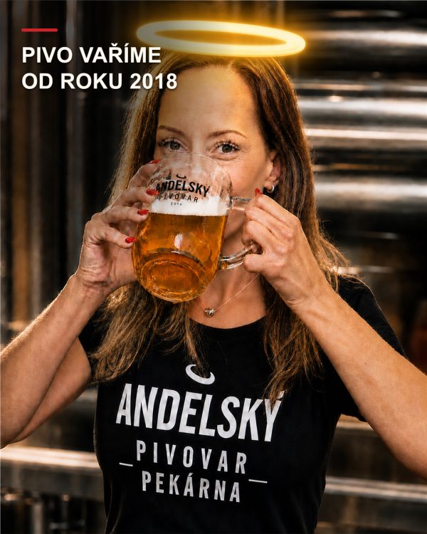 Členka týmu Andělského pivovaru drží značkový fasetovaný půllitr se světlým pivem před nerezovým vybavením pivovaru.